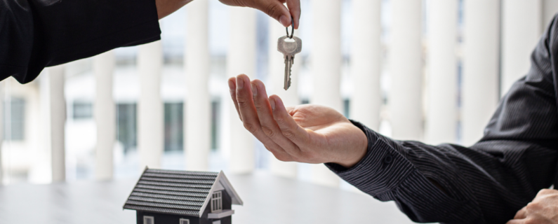 Property Management A hand reaches out to pass a key to another open hand over a table with a small house model in the background